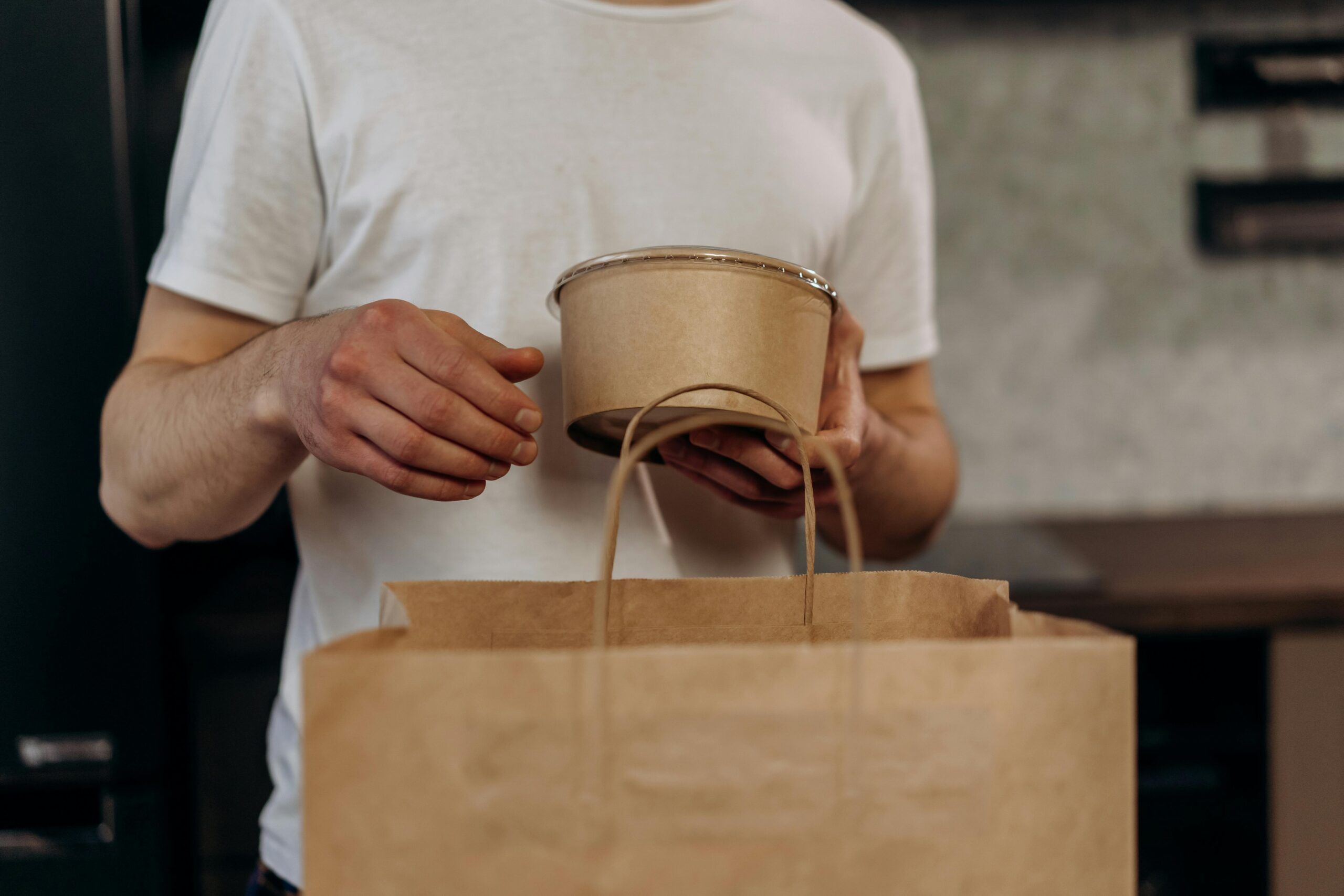 Close-up of a person holding a biodegradable container and paper bag indoors.